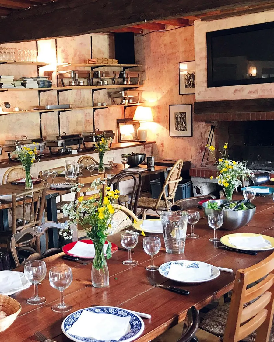 La salle à manger : longues tables dressées, bouquets de fleurs des champs, grande cheminée